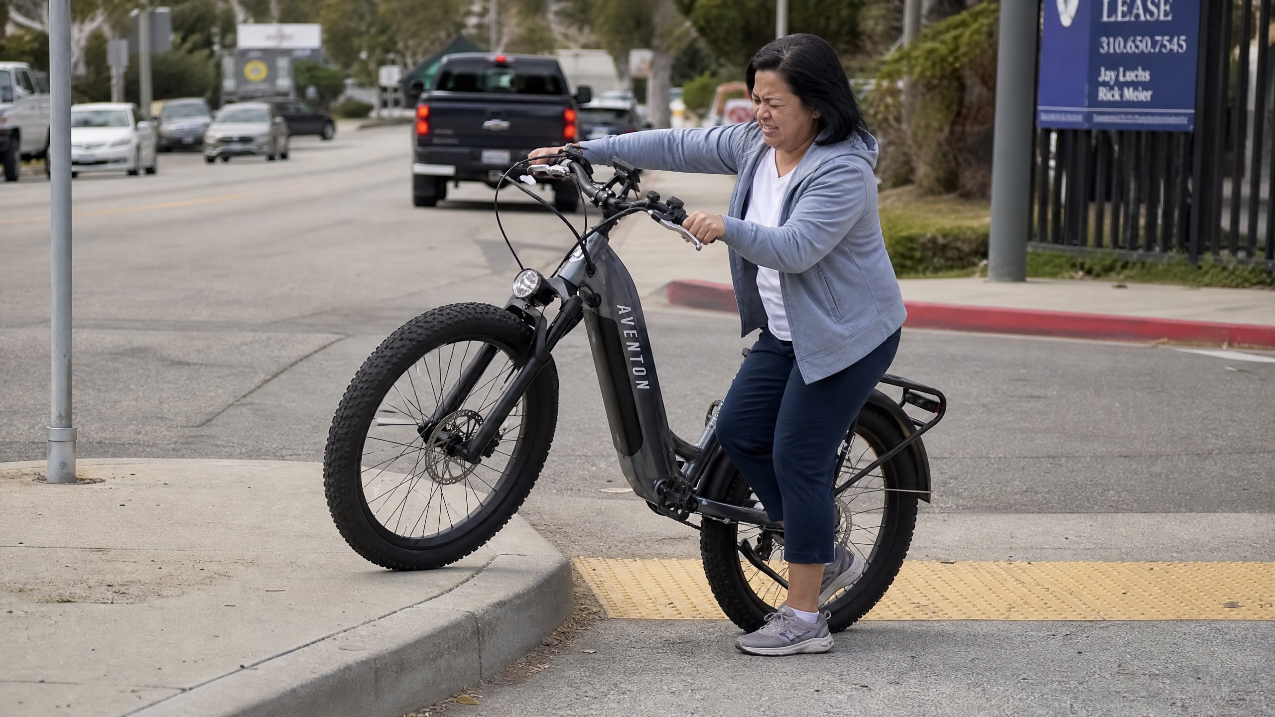 A short woman struggling to maneuver a heavy electric bike at low speed or trying to lift its front wheel over a curb, emphasizing the weight issue.